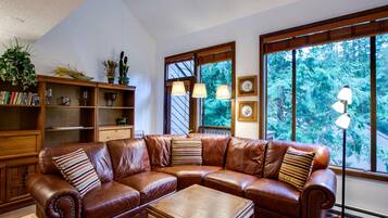 A brown leather sectional sofa with a wooden coffee table in a room with large windows, a bookshelf, and a floor lamp.