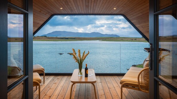 A wooden deck with a table and chairs, overlooking a lake with mountains in the background. - Ardcana (Strath)