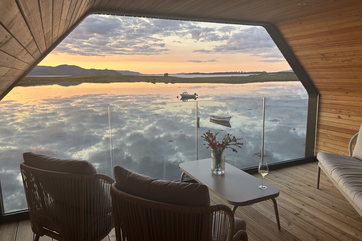 Una habitación con un gran ventanal que da a un cuerpo de agua y montañas distantes, con sillas de mimbre y una mesa con un florero y un vaso.