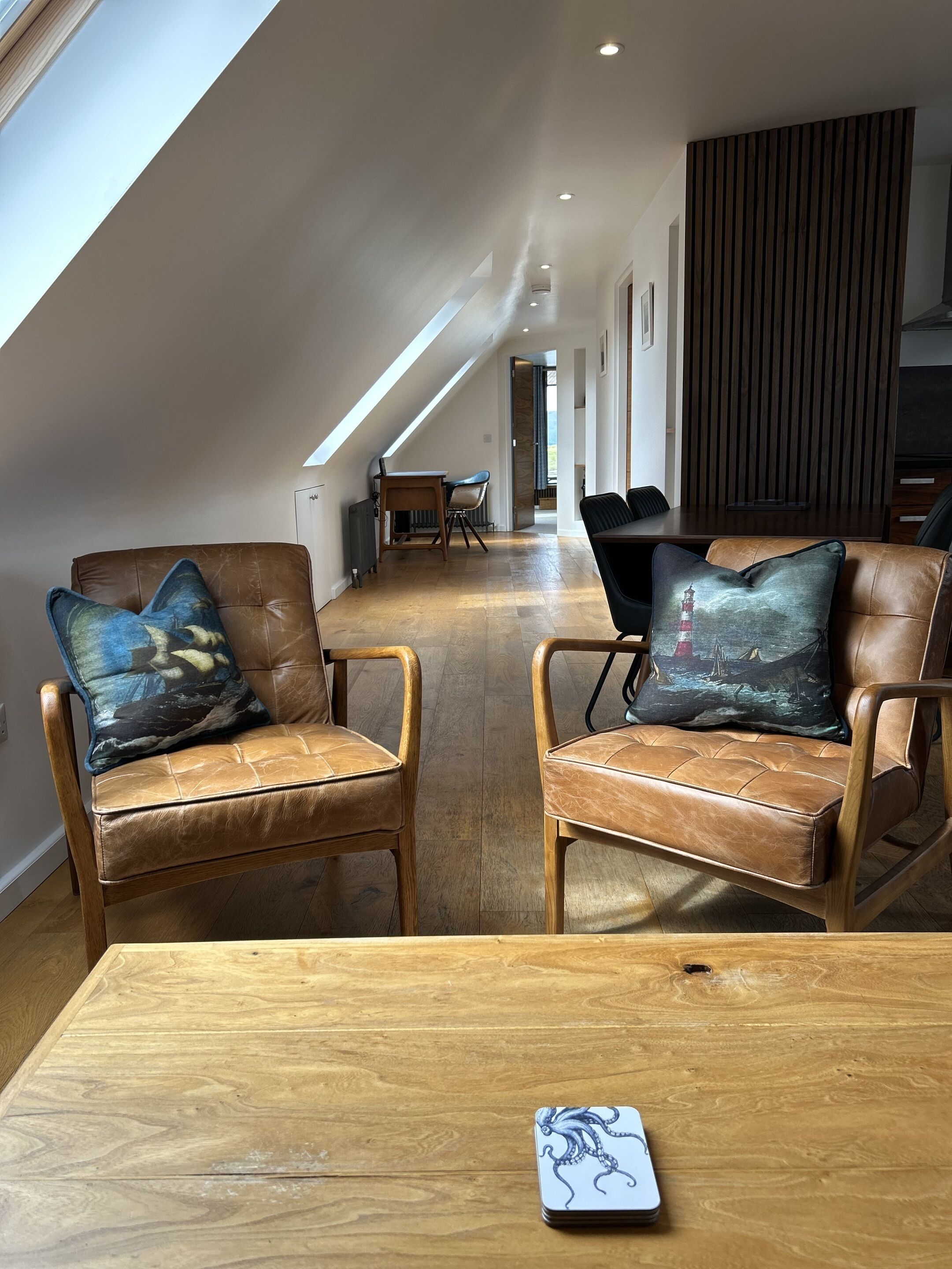 A spacious loft with high ceilings, a wooden dining table, leather armchairs with decorative pillows, and a view through an open door.