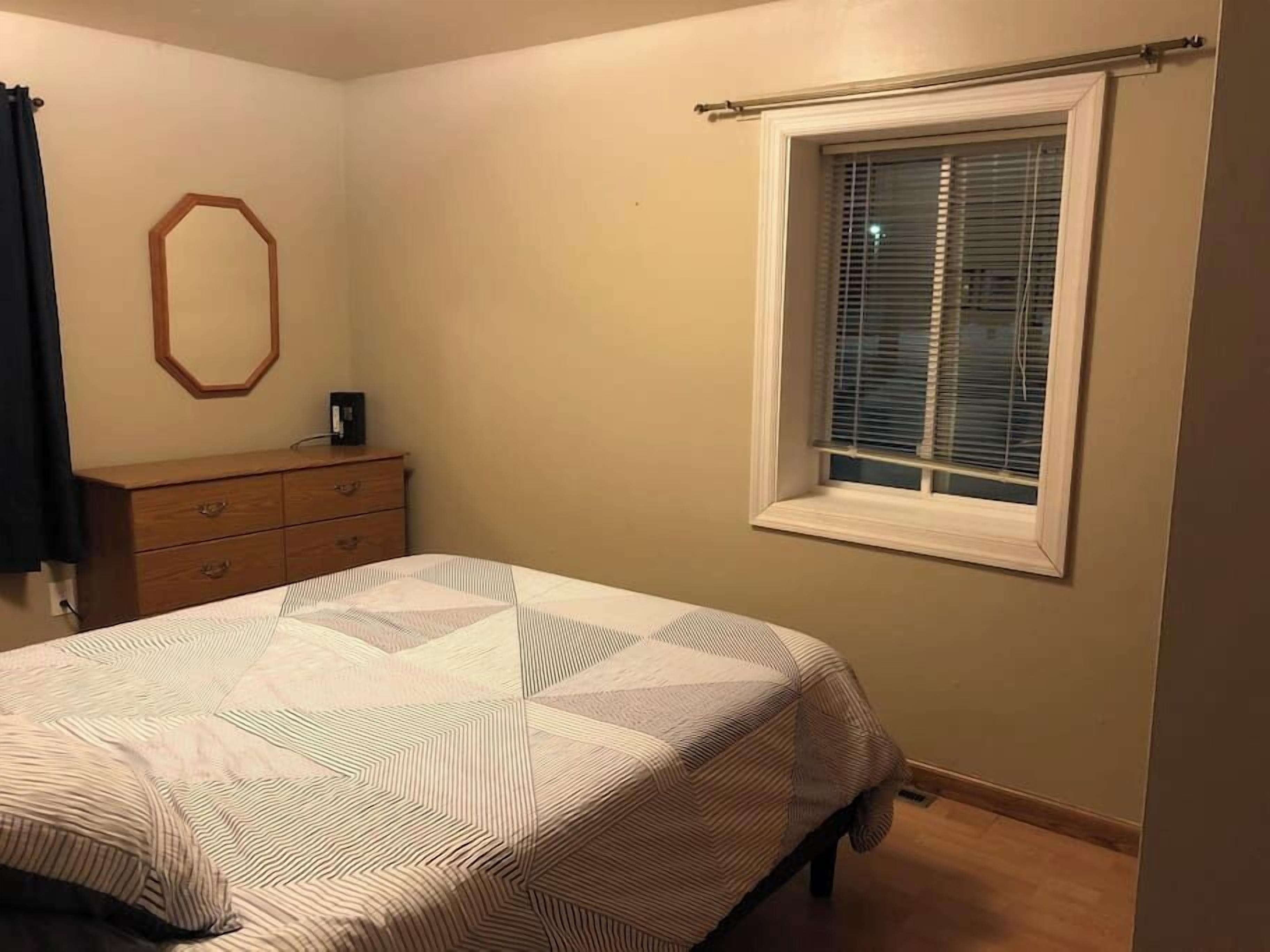 A bedroom with a bed, a dresser, a window with blinds, and a wall-mounted mirror.