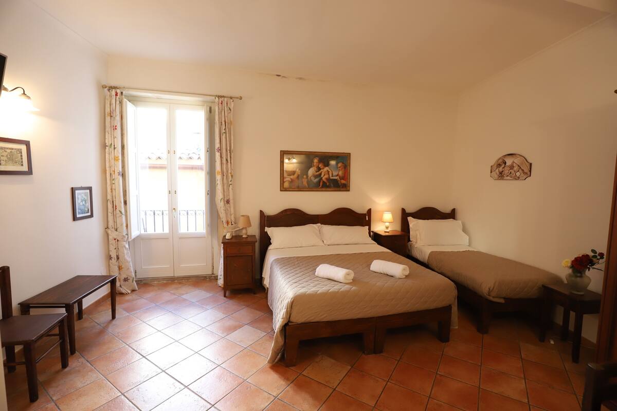 A bedroom with a double bed, a single bed, a desk, and a chair. There is a window with curtains and a painting on the wall.