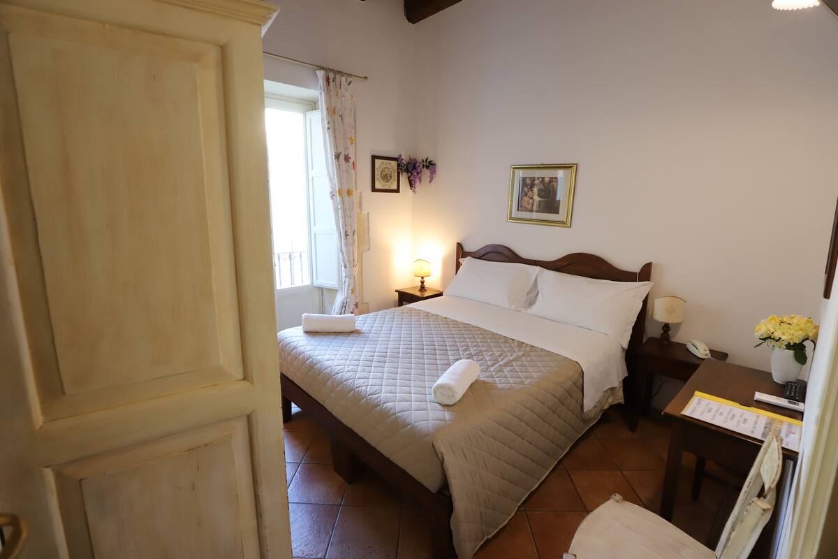 A hotel room with a bed, bedside table, lamp, and a window with curtains.