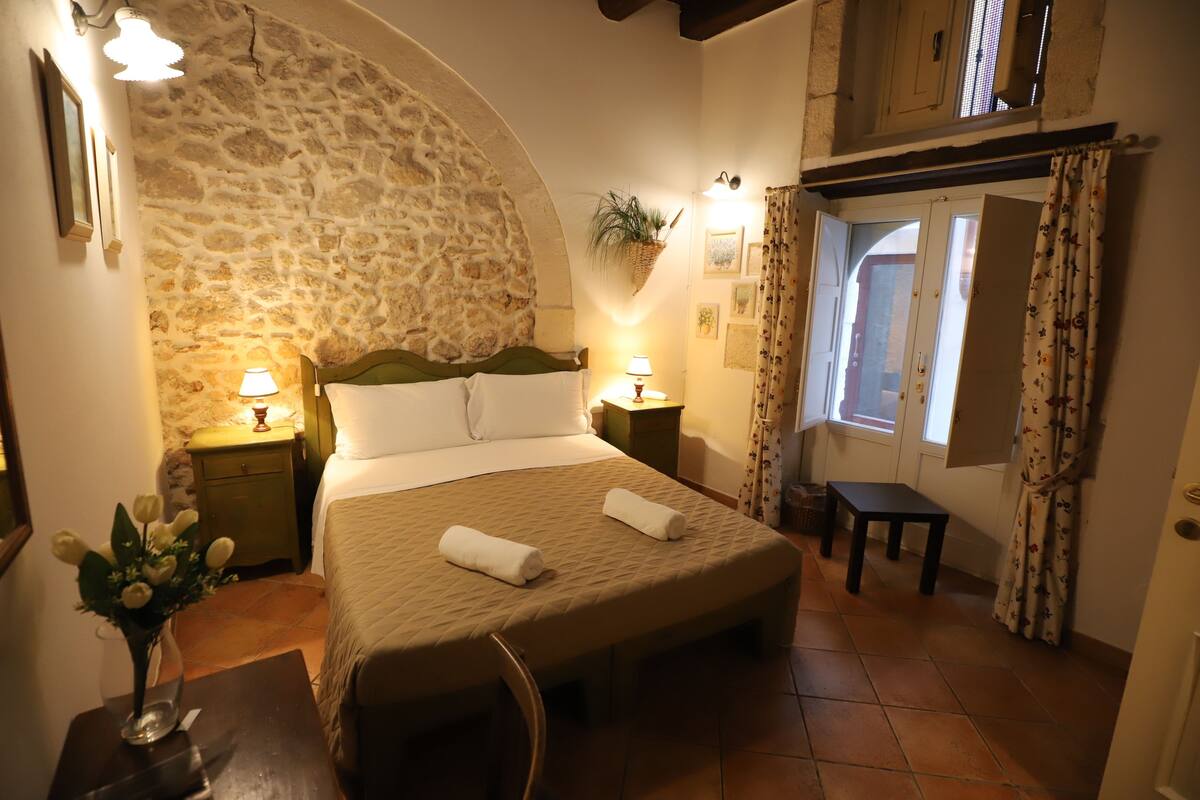 A cozy bedroom with a stone wall, a bed with two pillows, a bedside table with a lamp, a small table, and a window with curtains.