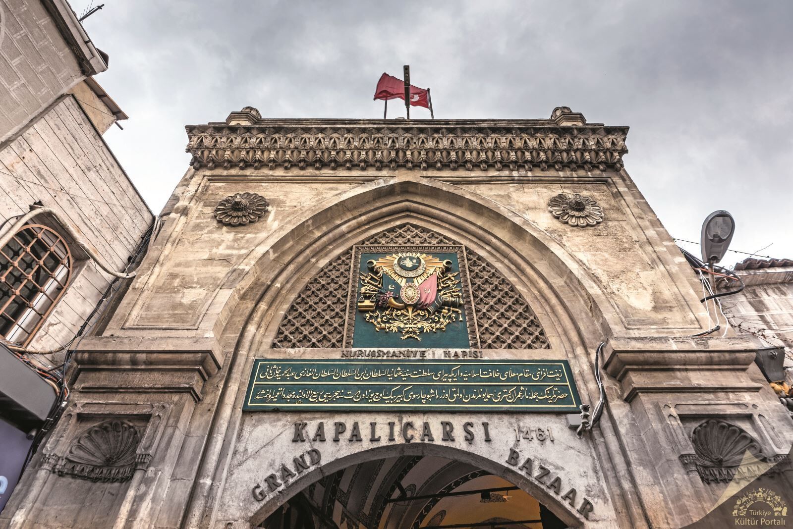 A historic stone archway with intricate carvings and a coat of arms, leading to a grand bazaar.