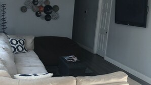 Ett vardagsrum med en beige soffa, ett soffbord, en väggmonterad TV och en dekorativ väggdekoration.