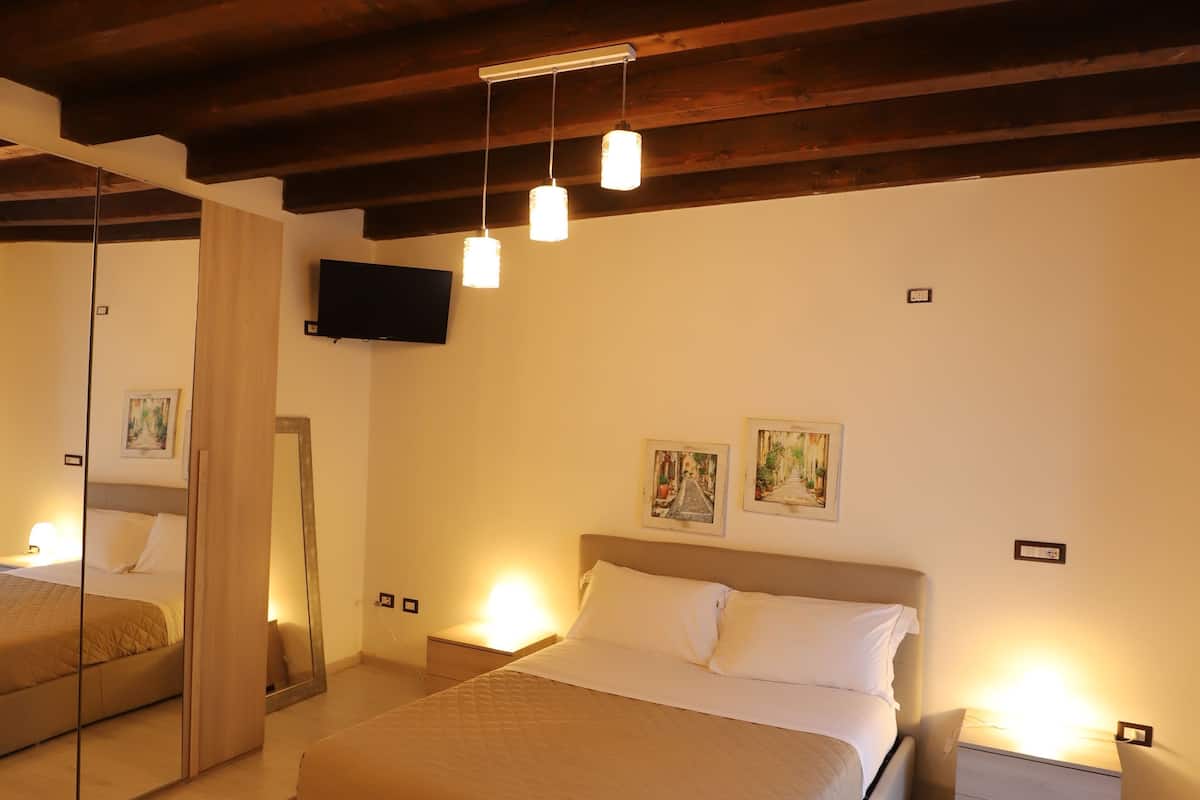 A bedroom with a large bed, wooden beams on the ceiling, a flat-screen TV, bedside lamps, and artwork on the wall.