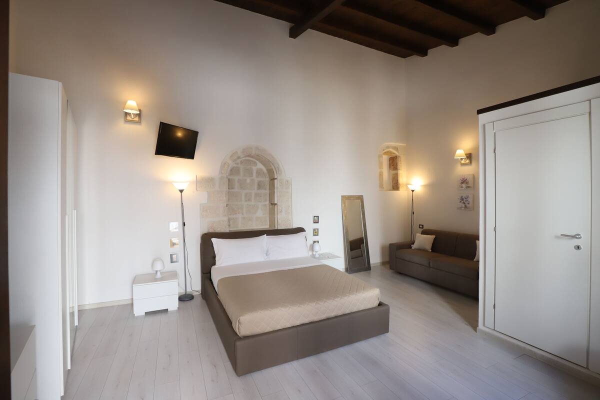 A bedroom with a bed, a nightstand, a TV mounted on the wall, a sofa, and a wardrobe.