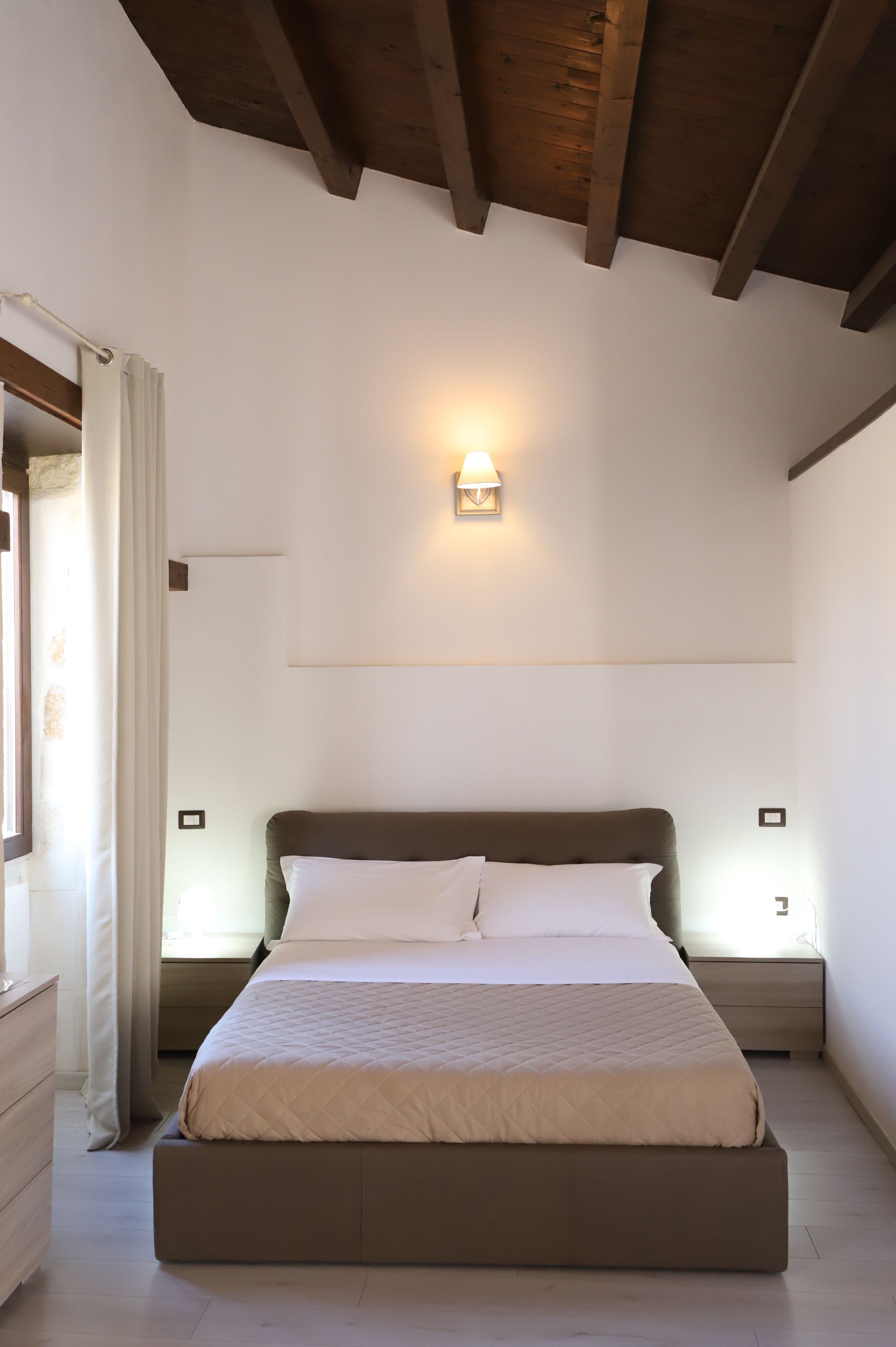 A bedroom with a bed, wooden ceiling beams, a wall-mounted light, and a window with curtains.