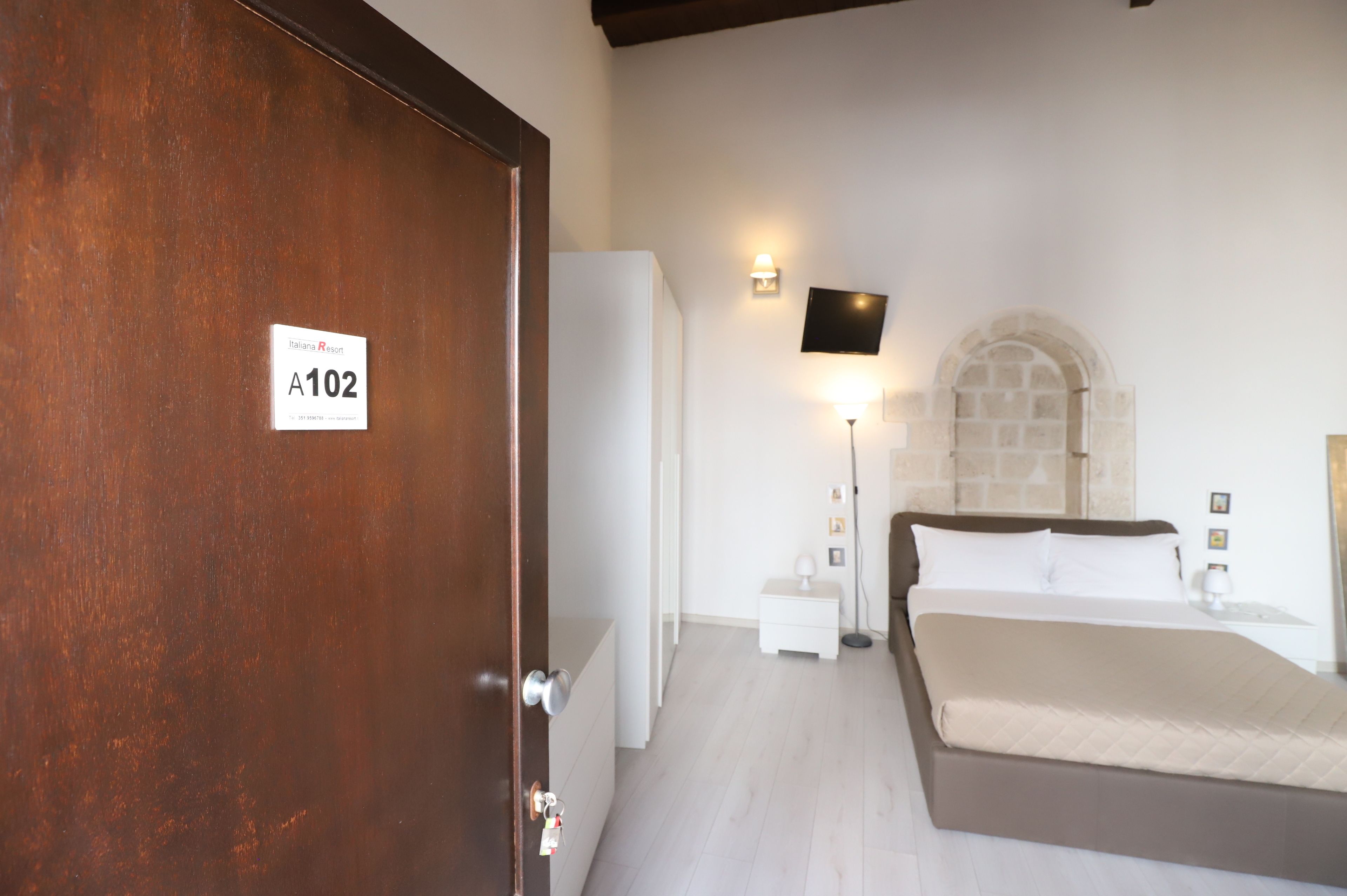 A hotel room with a wooden door labeled A102, a bed, a nightstand, a lamp, and a TV mounted on the wall.