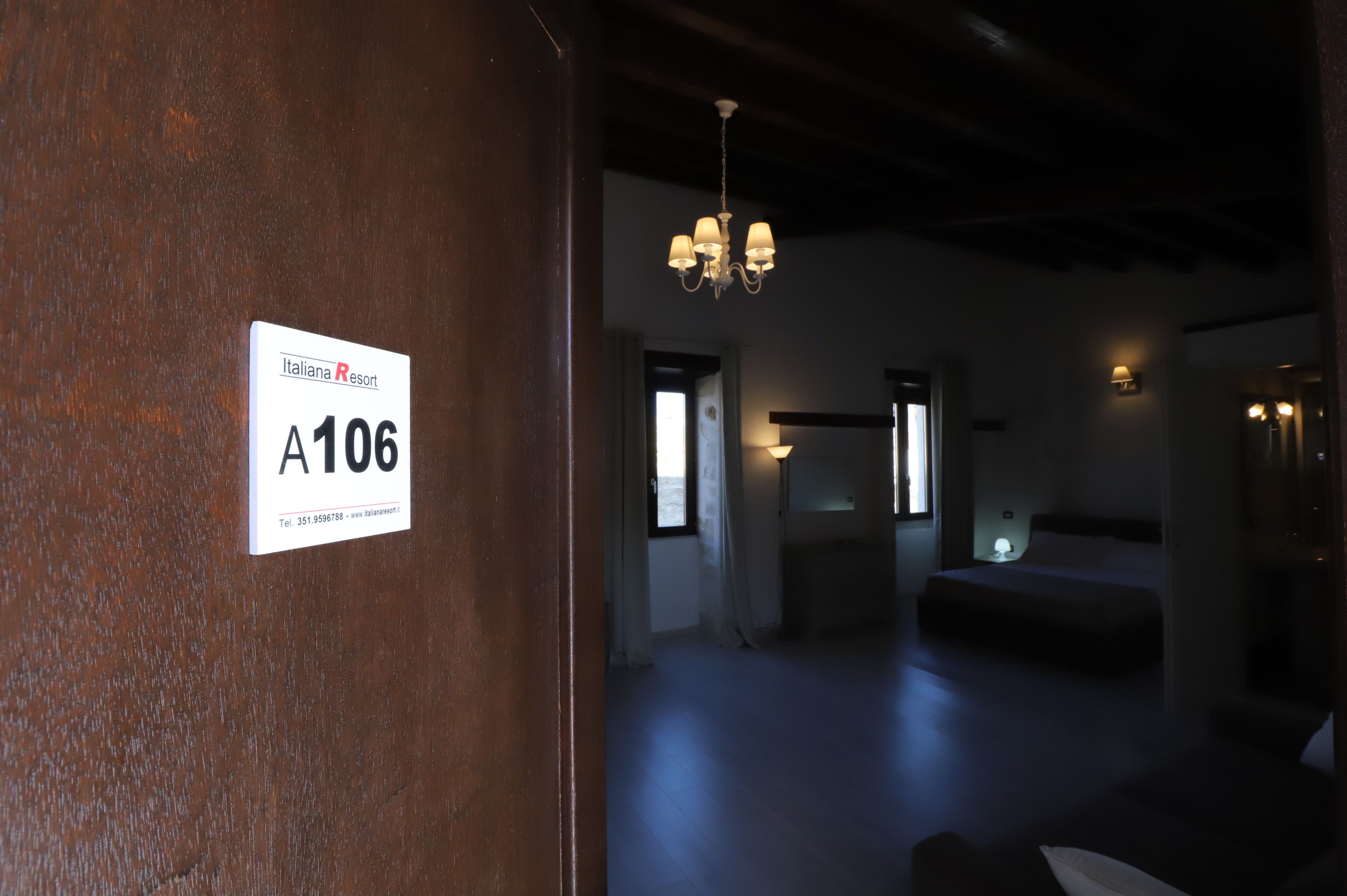 Une chambre d’hôtel avec un lit, un bureau et un lustre. Un panneau sur le mur indique le numéro de la chambre : A106.