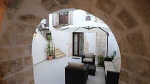 Un patio con suelo de baldosas blancas, un conjunto de sofá exterior negro, una sombrilla blanca y una planta en maceta.