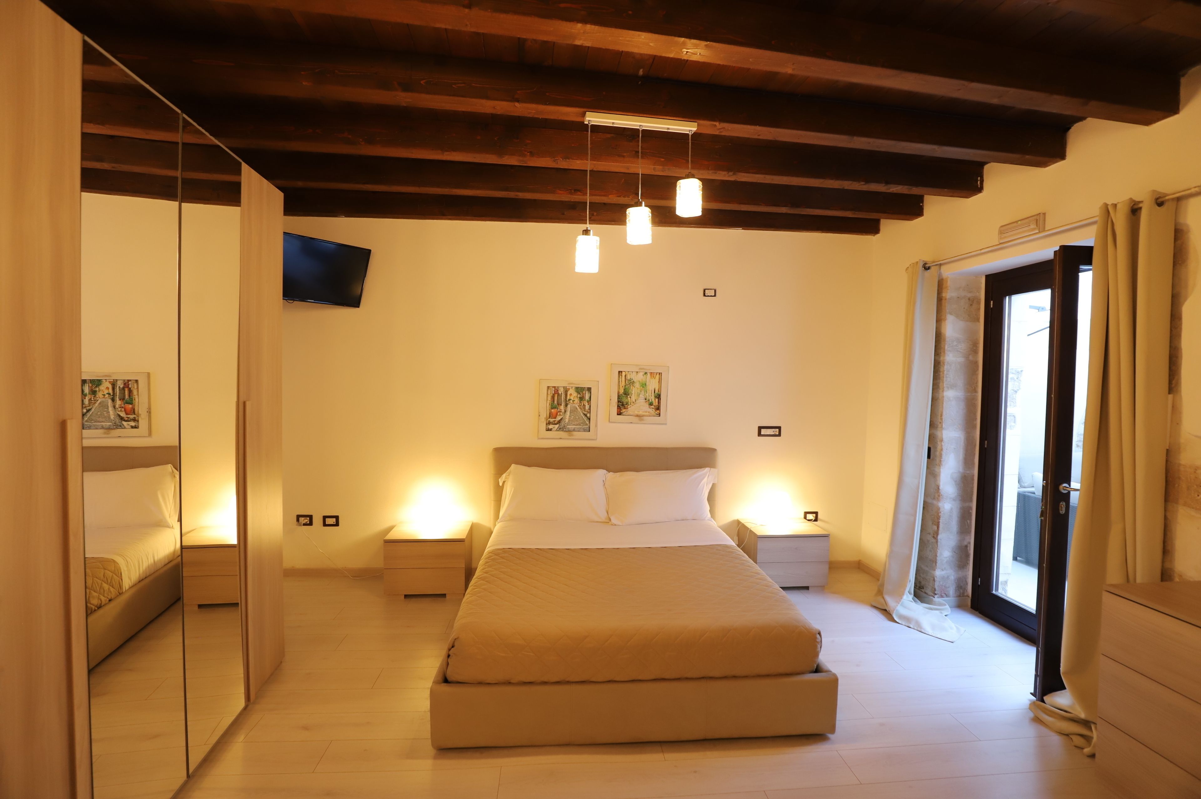 A bedroom with a large bed, bedside tables, a TV mounted on the wall, and a sliding glass door leading to a balcony.