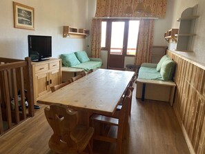 A room with a wooden dining table, benches, a sofa, a television, and a window with curtains.
