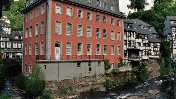 A red building with multiple windows and a balcony, adjacent to a river and surrounded by greenery.