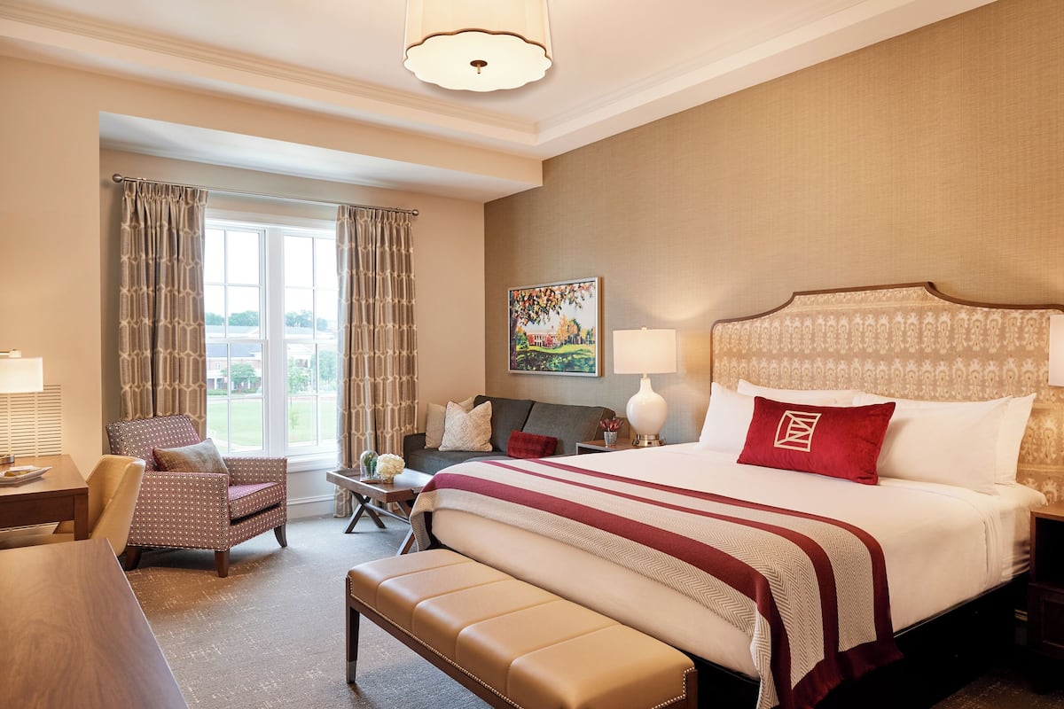 A hotel room with a large bed, a chair, a bench, a desk, and a window with curtains.