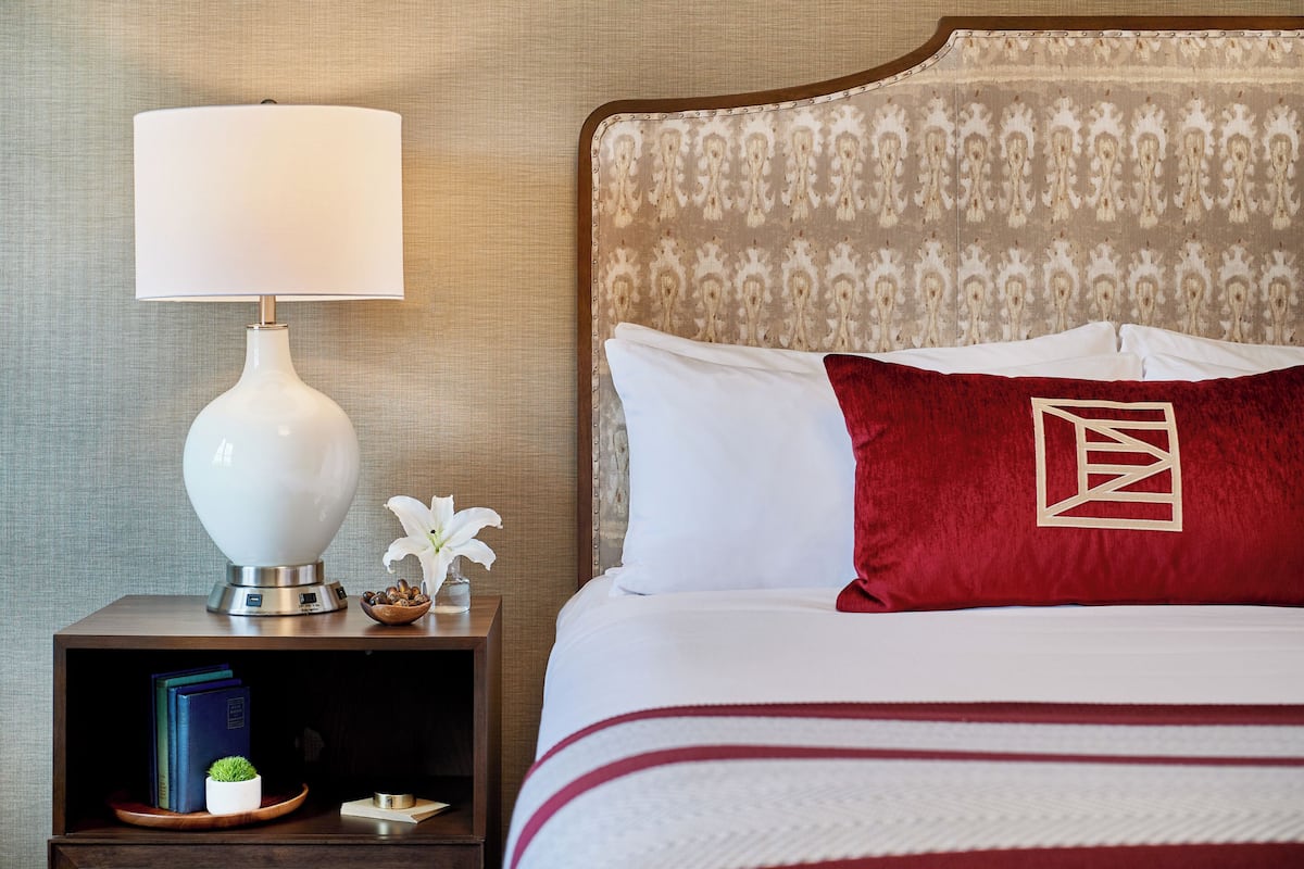 A neatly made bed with white and red bedding, a red pillow with a geometric design, a white lamp on a bedside table, and a small plant.