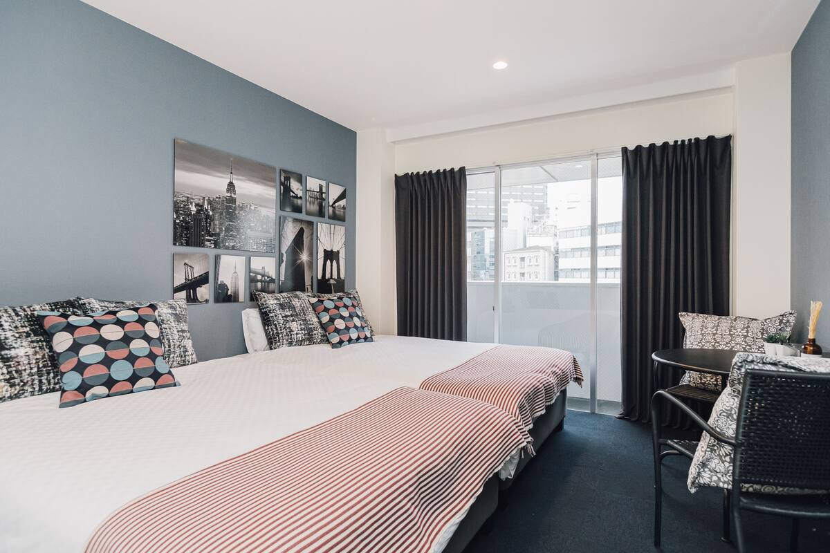 A modern hotel room with a large bed, a dining area, and a city view through the window.