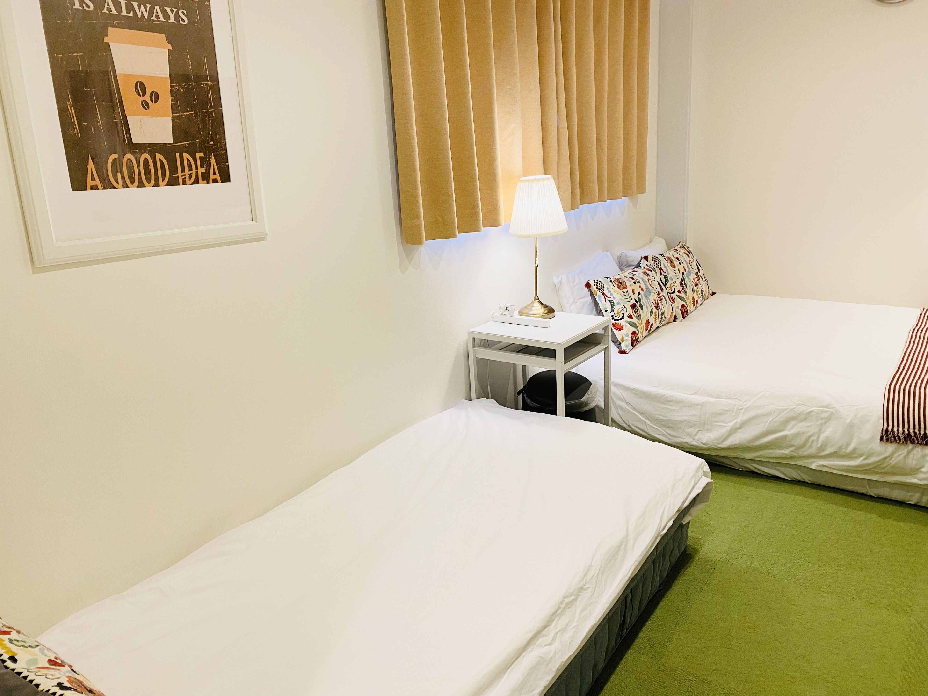 Ein Zimmer mit zwei Einzelbetten, einem kleinen Nachttisch mit Lampe, einem Poster an der Wand und einem Fenster mit Vorhängen.