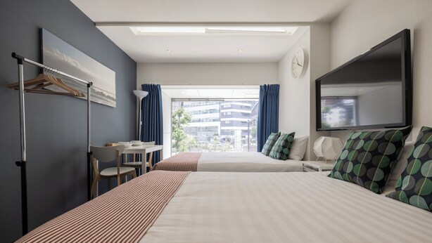 Habitación de hotel con dos camas, un escritorio, una silla, un televisor y vistas a edificios a través de la ventana.