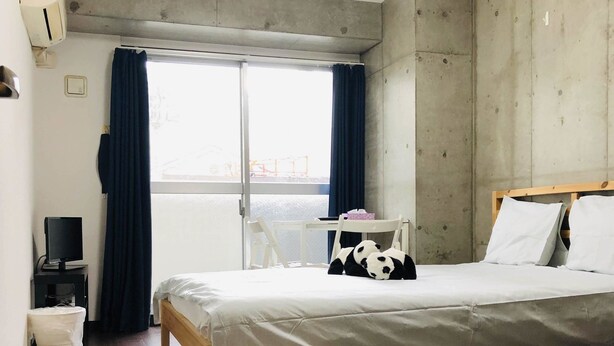 Een moderne slaapkamer met een houten vloer, een bed met wit beddengoed, een klein nachtkastje met een lamp, een raam met gordijnen en een betonnen muur.