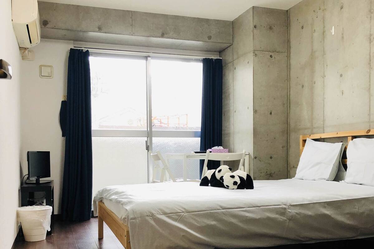 A modern bedroom with a wooden floor, a bed with white bedding, a small bedside table with a lamp, a window with curtains, and a concrete wall.