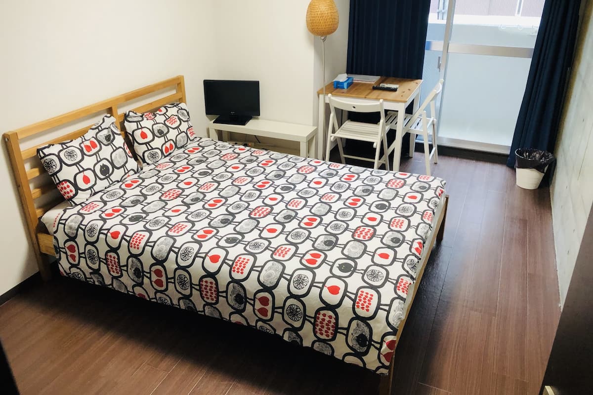A neatly made bed with a patterned bedspread, a small desk with a chair, a lamp, and a window with curtains.