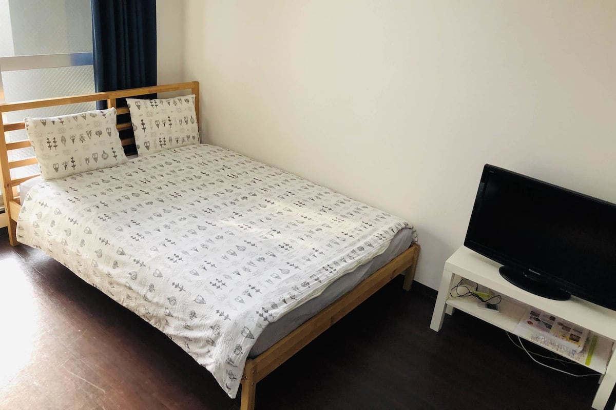 A single bed with a patterned bedspread, a wooden headboard, and matching pillows. A flat-screen TV is on a white stand to the right.