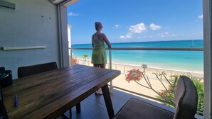 A room with a view of a beach, a dining table, and a person standing by the window.