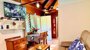 A living room with a TV, wooden furniture, a sofa with patterned cushions, and a dining area with a table and chairs.