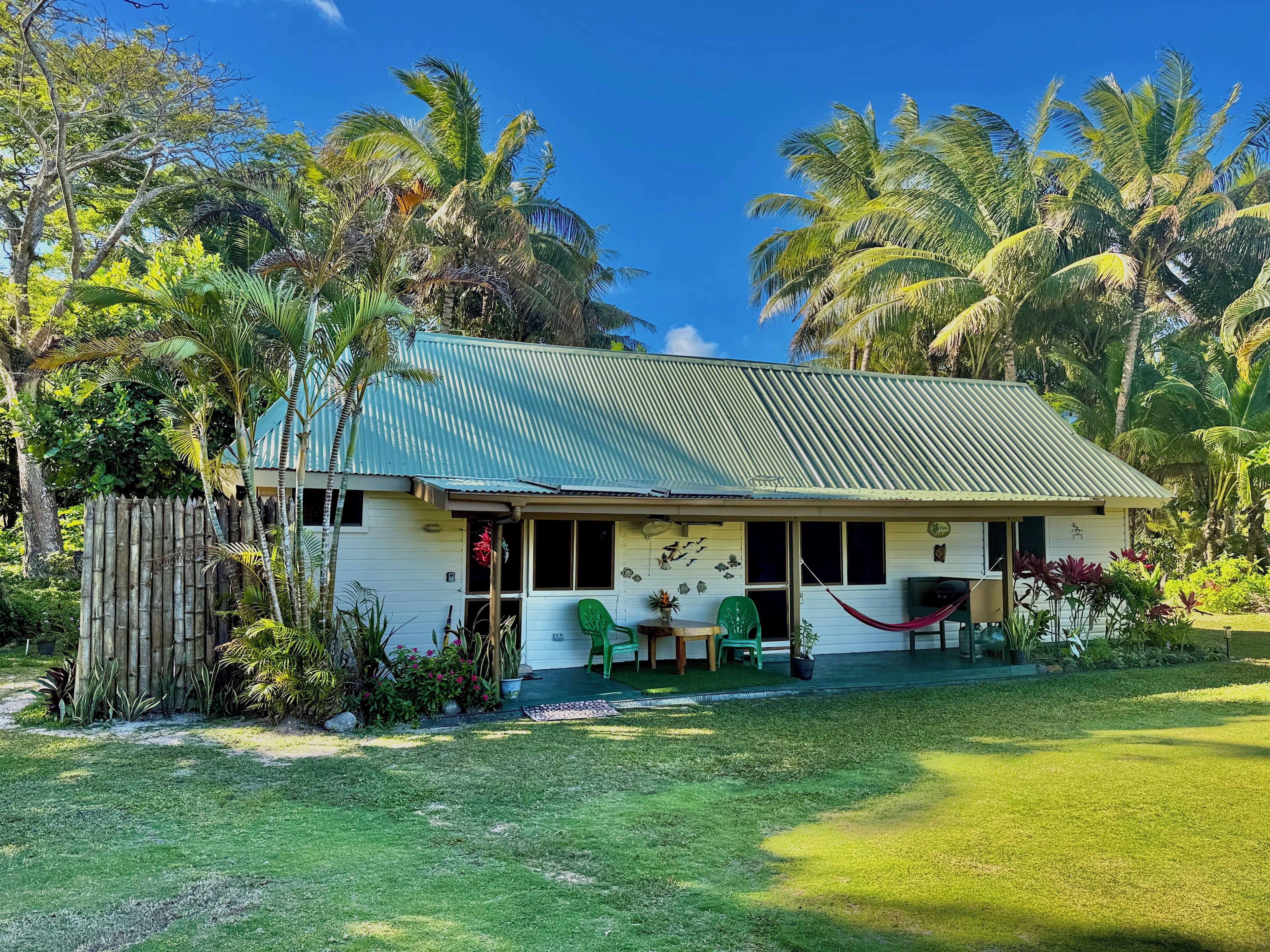 A single-story house with a corrugated metal roof, surrounded by lush greenery and palm trees.
