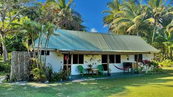 A single-story house with a corrugated metal roof, surrounded by lush greenery and palm trees.