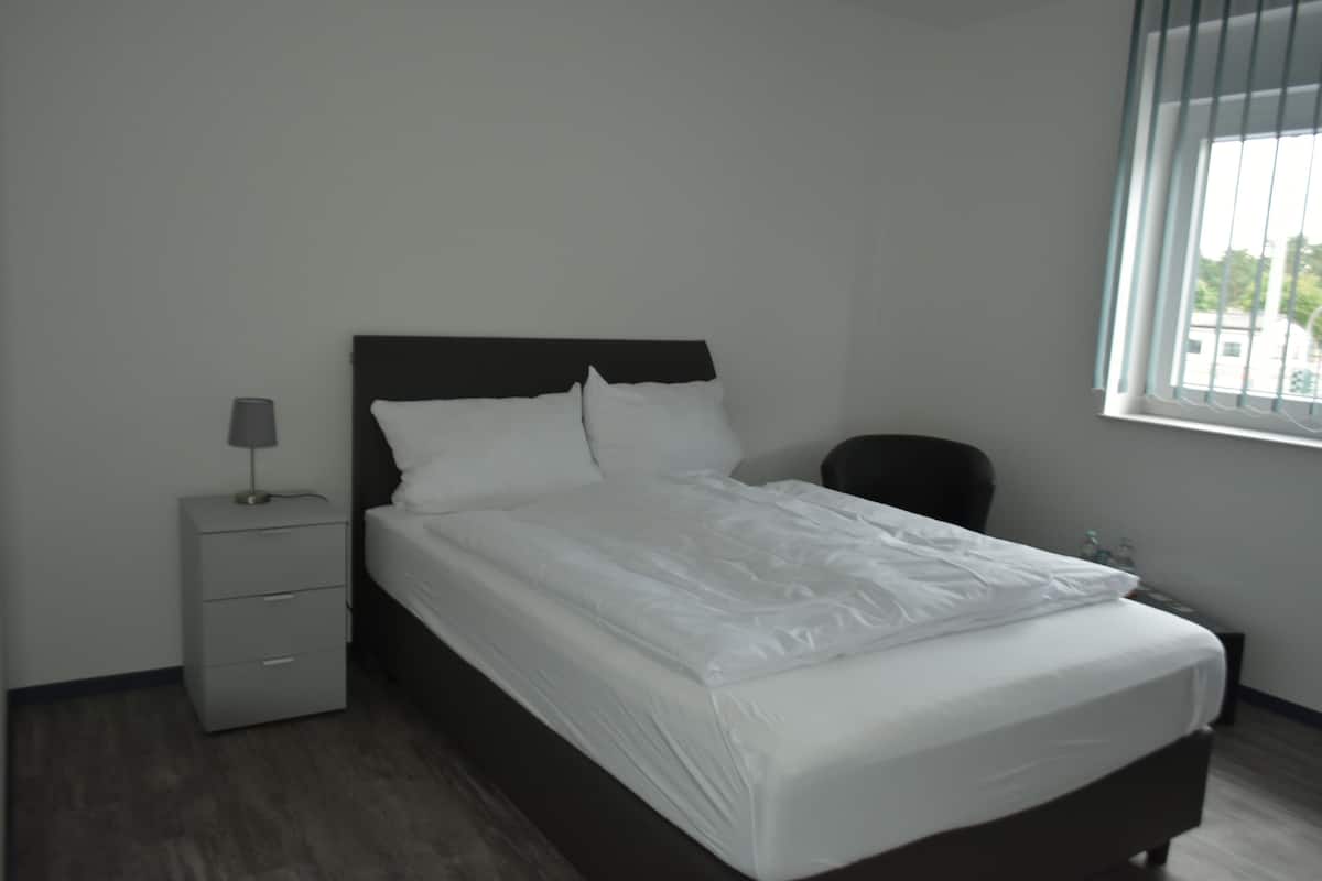 Een net opgemaakt bed met witte lakens in een eenvoudige, moderne kamer met een houten vloer en een klein nachtkastje met een lamp.