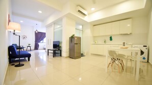 A modern living room with a sofa, a table, and a TV. There is a kitchen area with a refrigerator, a washing machine, and cabinets. - GT Home Encorp Strand Residence (Petaling Jaya)