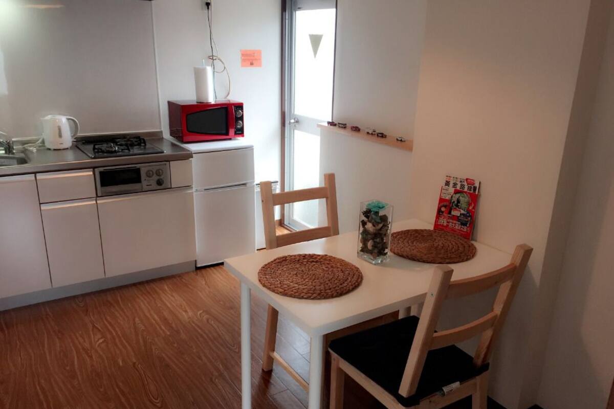 A compact kitchen with a microwave, kettle, and gas stove. There is a dining table with two chairs and a small decorative item on it.