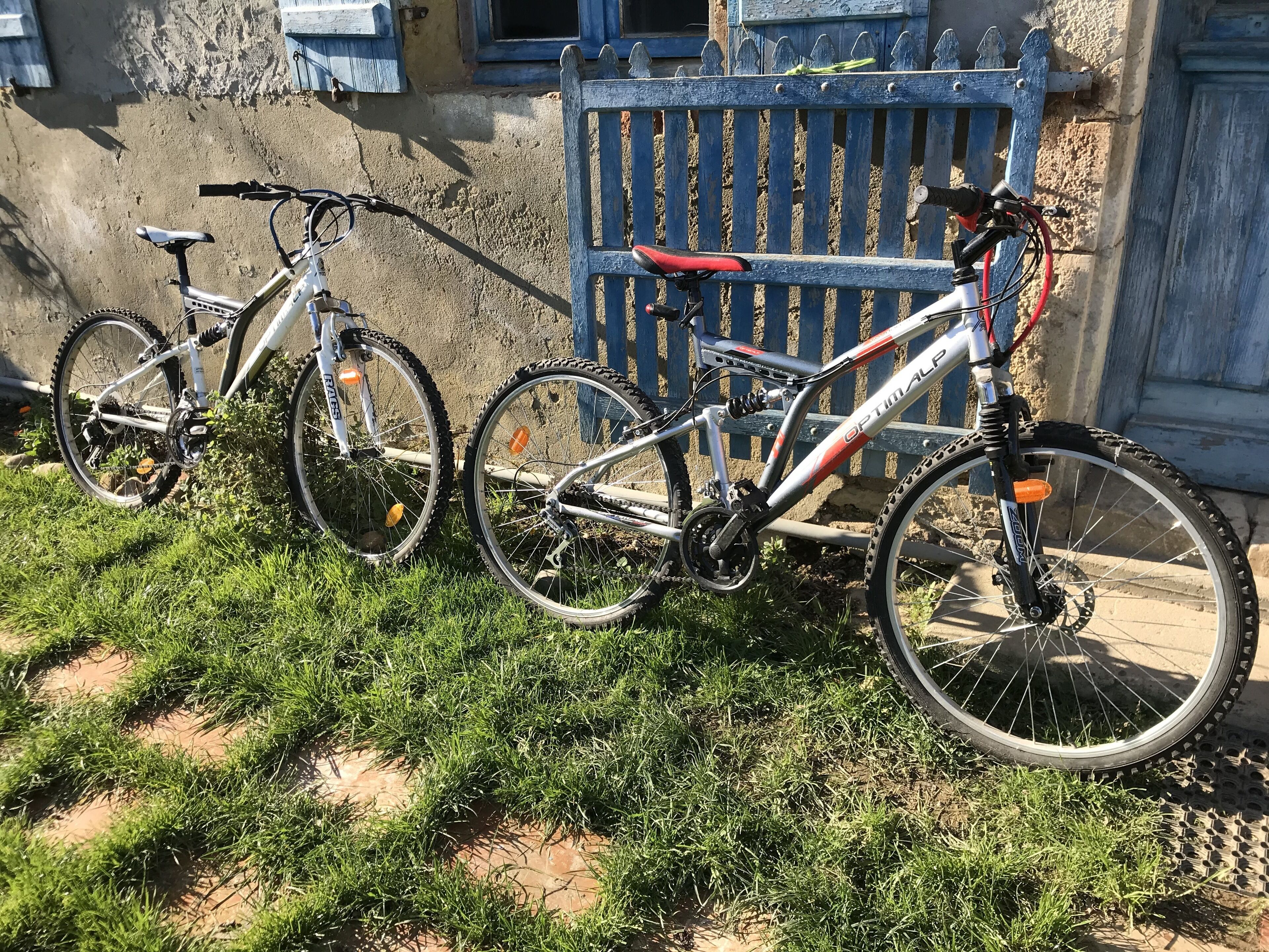 Two bicycles parked on a grassy area in front of a building with a blue fence and a window with blue shutters.