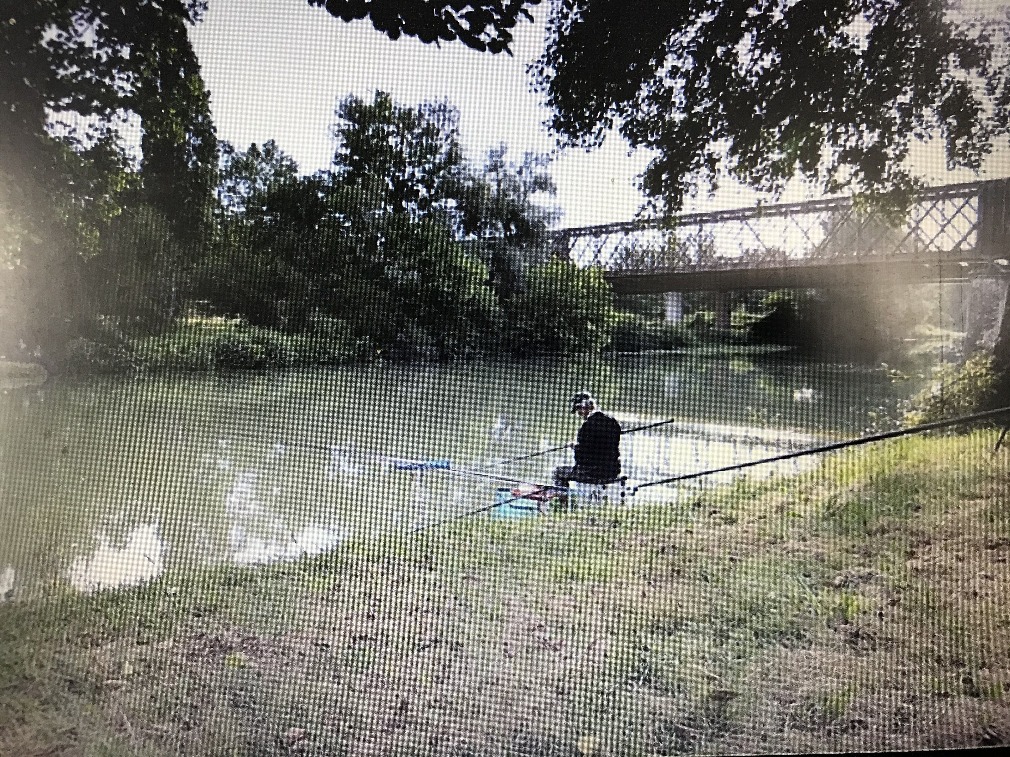 A person fishing by a calm river with a bridge in the background.
