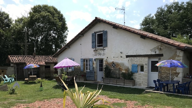 A rustic house with a tiled roof, a stone wall, and a wooden deck. There are two outdoor tables with chairs and a purple umbrella. - Chambres Bambou & Corail & Music (Saint Sever)