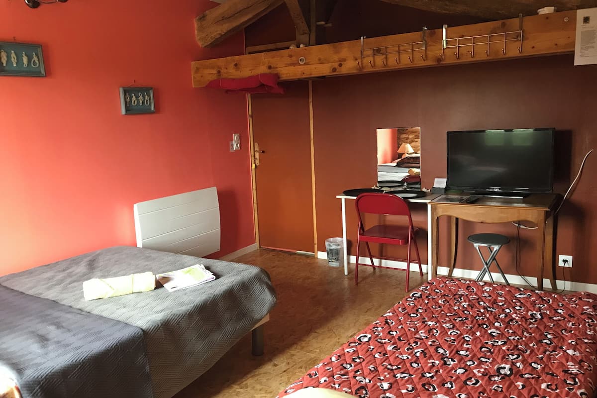 A room with two beds, a desk with a computer and TV, a chair, and a wooden ceiling structure.