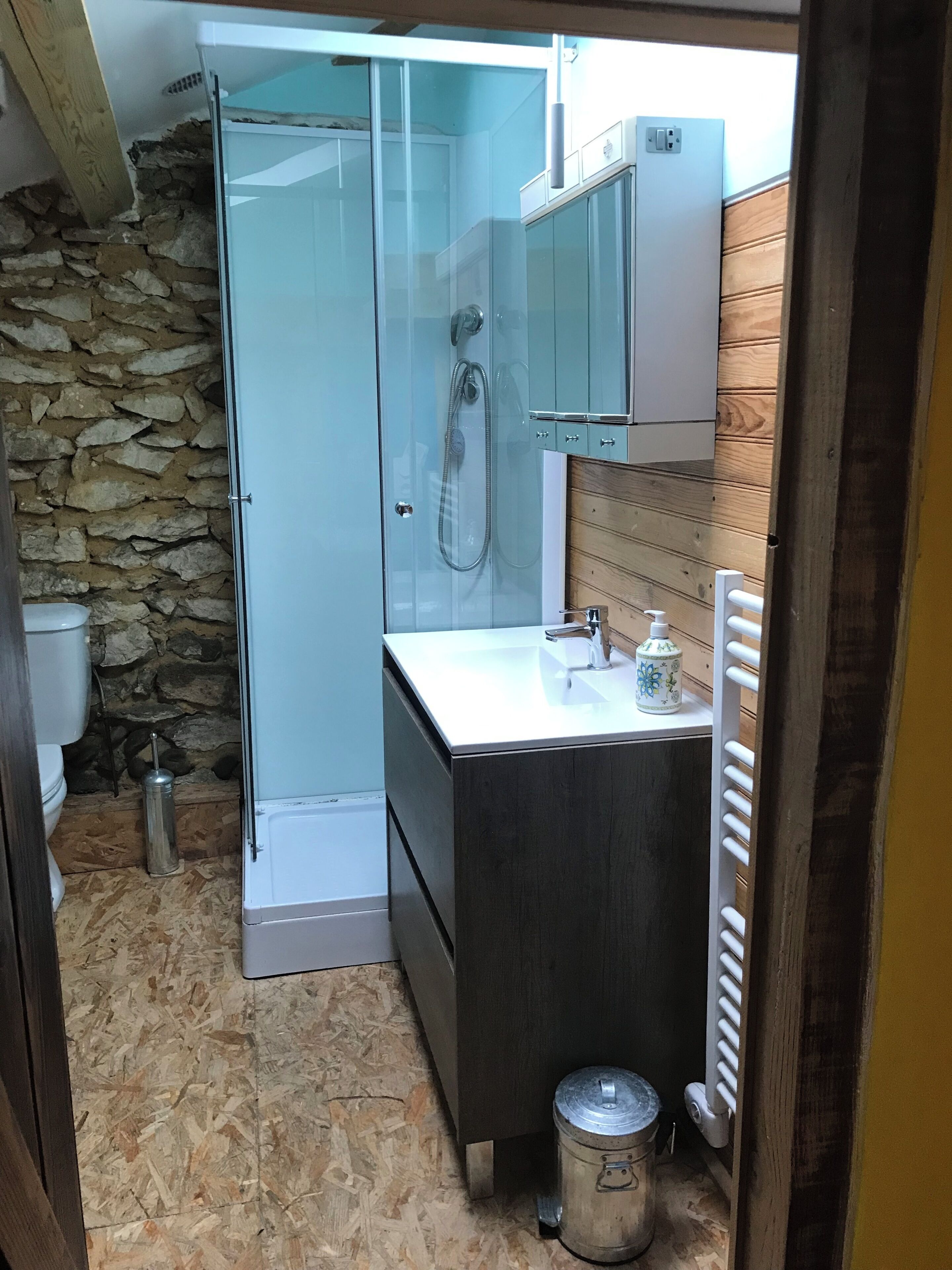 Casa de banho compacta com cabine de duche em vidro, lavatório branco com torneira, pequeno recipiente de porcelana e radiador.