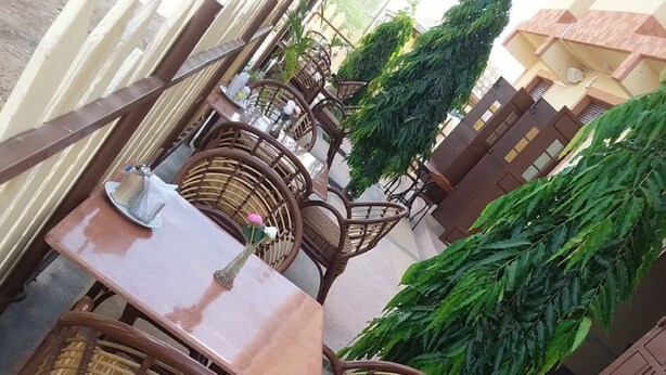 An outdoor seating area with wicker chairs and tables, surrounded by potted plants and a building with a balcony.