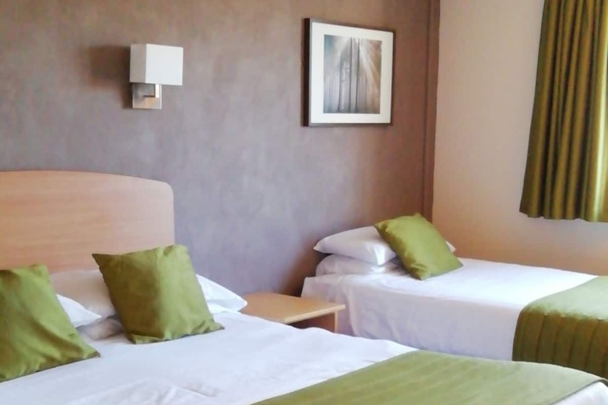 Une chambre d’hôtel avec deux lits, chacun agrémenté de draps verts et d’oreillers blancs. Un luminaire mural et un tableau encadré sont fixés au mur, au-dessus des lits.
