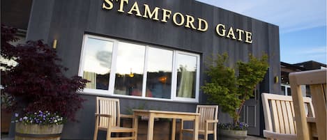 A modern building with the name "STAMFORD GATE" prominently displayed. There is a wooden table and chairs set outside, and two large planters with flowers.