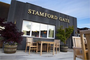 A modern building with the name "STAMFORD GATE" prominently displayed. There is a wooden table and chairs set outside, and two large planters with flowers.
