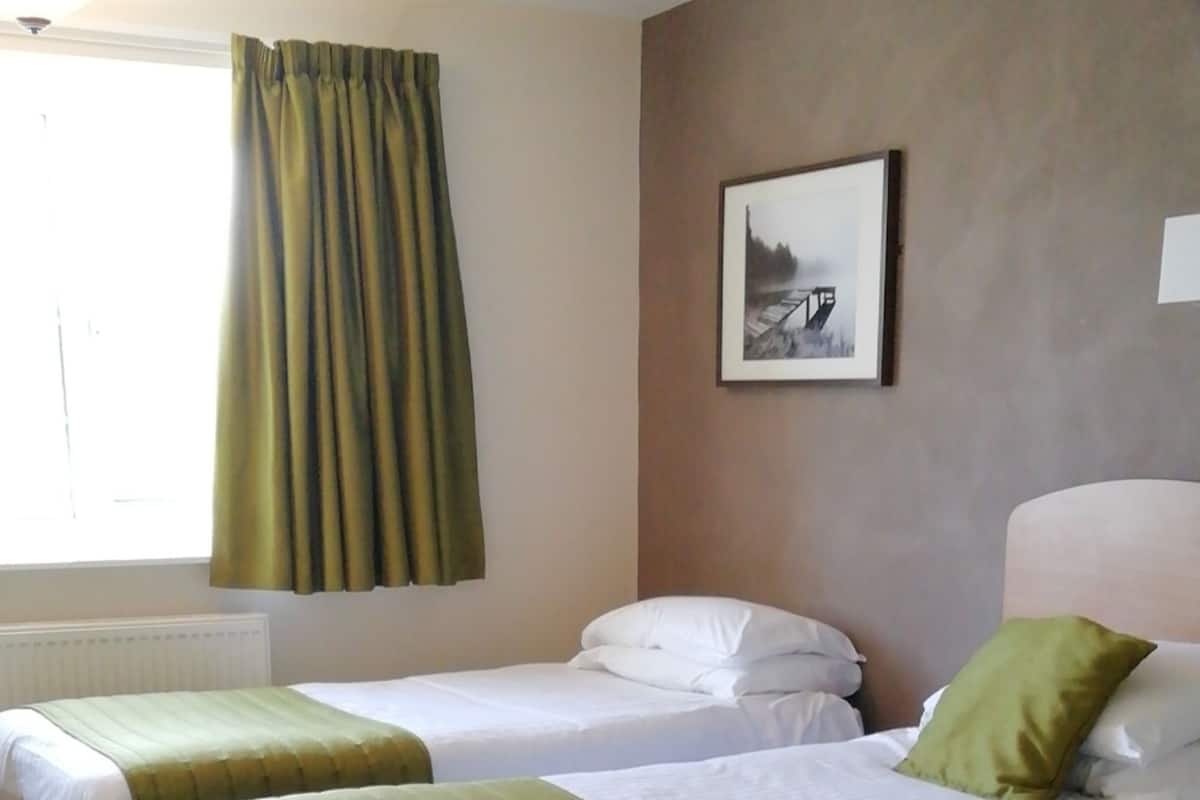Une chambre d’hôtel avec deux lits simples, chacun recouvert d’un plaid vert et de draps blancs. Il y a une fenêtre avec des rideaux verts, un tableau encadré au mur et un radiateur sous la fenêtre.