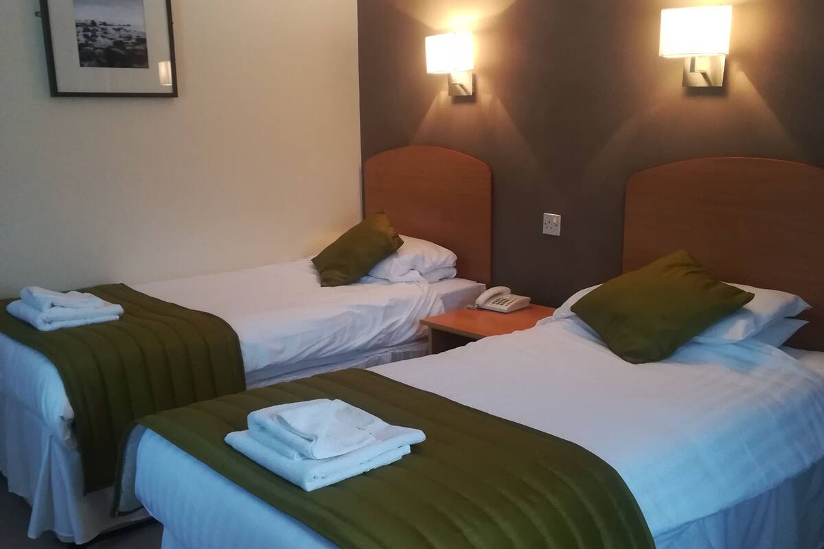 Une chambre d’hôtel avec deux lits, chacun recouvert d’une couvre-lit verte et blanche, une table de chevet avec un téléphone et des lampes fixées au mur.