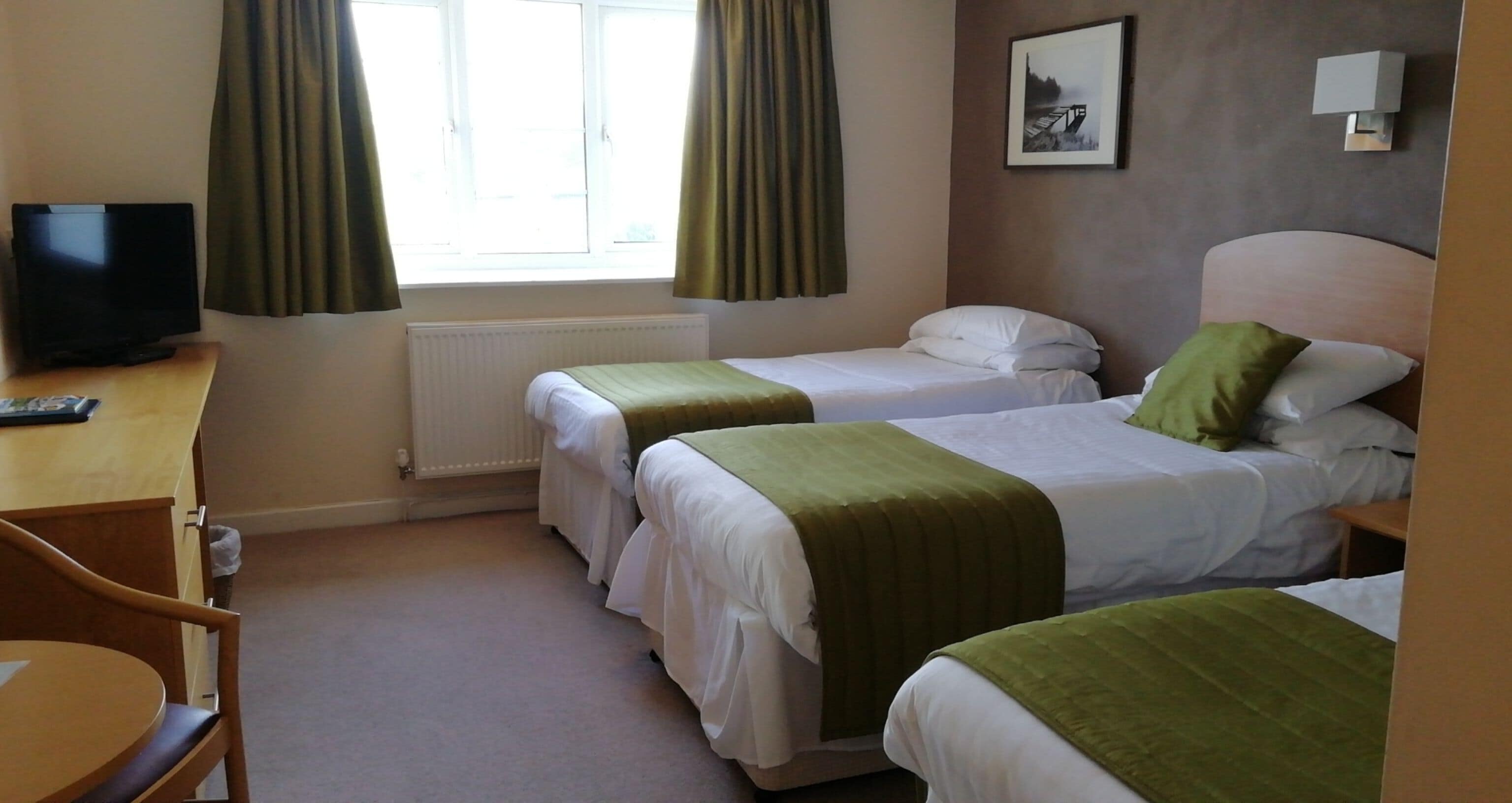 A hotel room with two single beds, a desk with a television, a radiator, and a window with curtains.