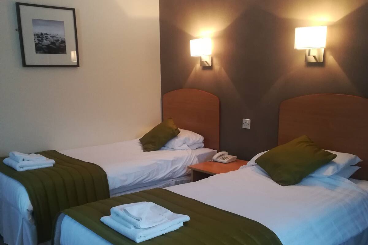 Une chambre d’hôtel avec deux lits, chacun recouvert d’une couverture verte, doté de draps blancs et muni d’un téléphone sur la table de chevet.