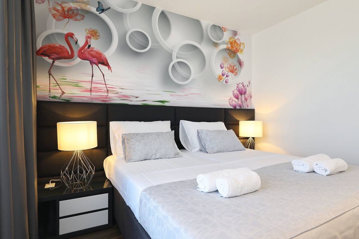 Una camera d'albergo con un letto grande, due comodini con lampade, una parete decorata con motivi floreali e uccelli, e asciugamani ordinatamente piegati sul letto.