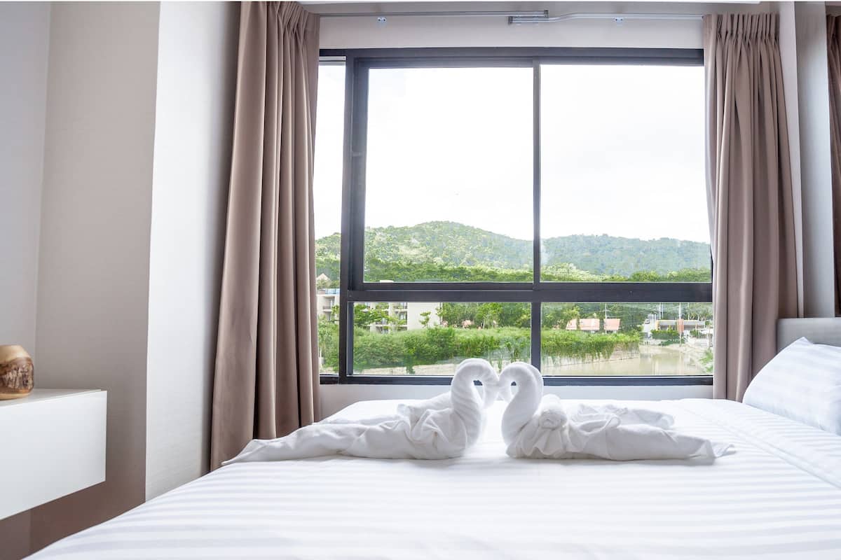 Een hotelkamer met een groot raam, een bed met witte lakens en uitzicht op groen en gebouwen.